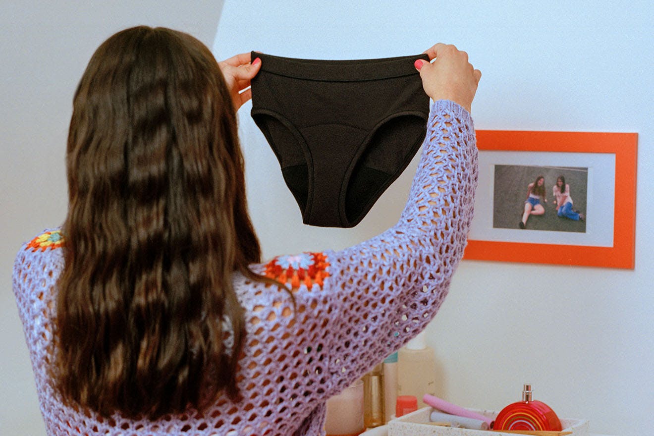 Teen girl facing backwards holding up a pair of Thinx Teens Bikini reusable period underwear in the color black. 