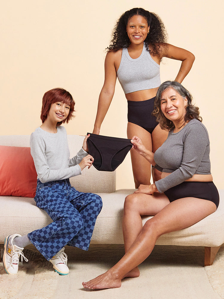 three models on a couch holding a pair of black period underwear