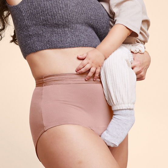 A mother stands smiling in front of a wooden crib and a beige background, wearing a pink shirt and pair of Thinx period underwear in the style black Hi-Waist, holding her baby who is wearing a yellow bodysuit and wiping the baby’s mouth with a cloth